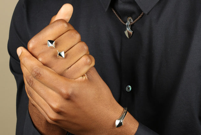 Two mens hands being held together in a clasp. The man is wearing a silver Arrowhead ring, a silver arrowhead bangle and an arrowhead necklace on leather. He is wearing a blue navy button down shirt.